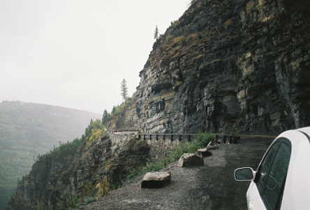 [Part of a white car is visible at the right edge of the photo. The large stones marking the edge of the pull-off become a guard rail just a short distance down the road. The rock face is near vertical and very segmented. The distance air and mountains are shrouded in fog. ]